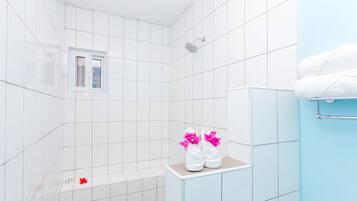 A modern bathroom with white tiles, a shower, a small window, and a shelf with towels and flowers.