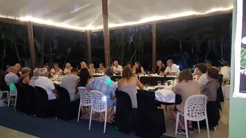 An outdoor dining area with round tables and white chairs, surrounded by a covered area with a view of palm trees.