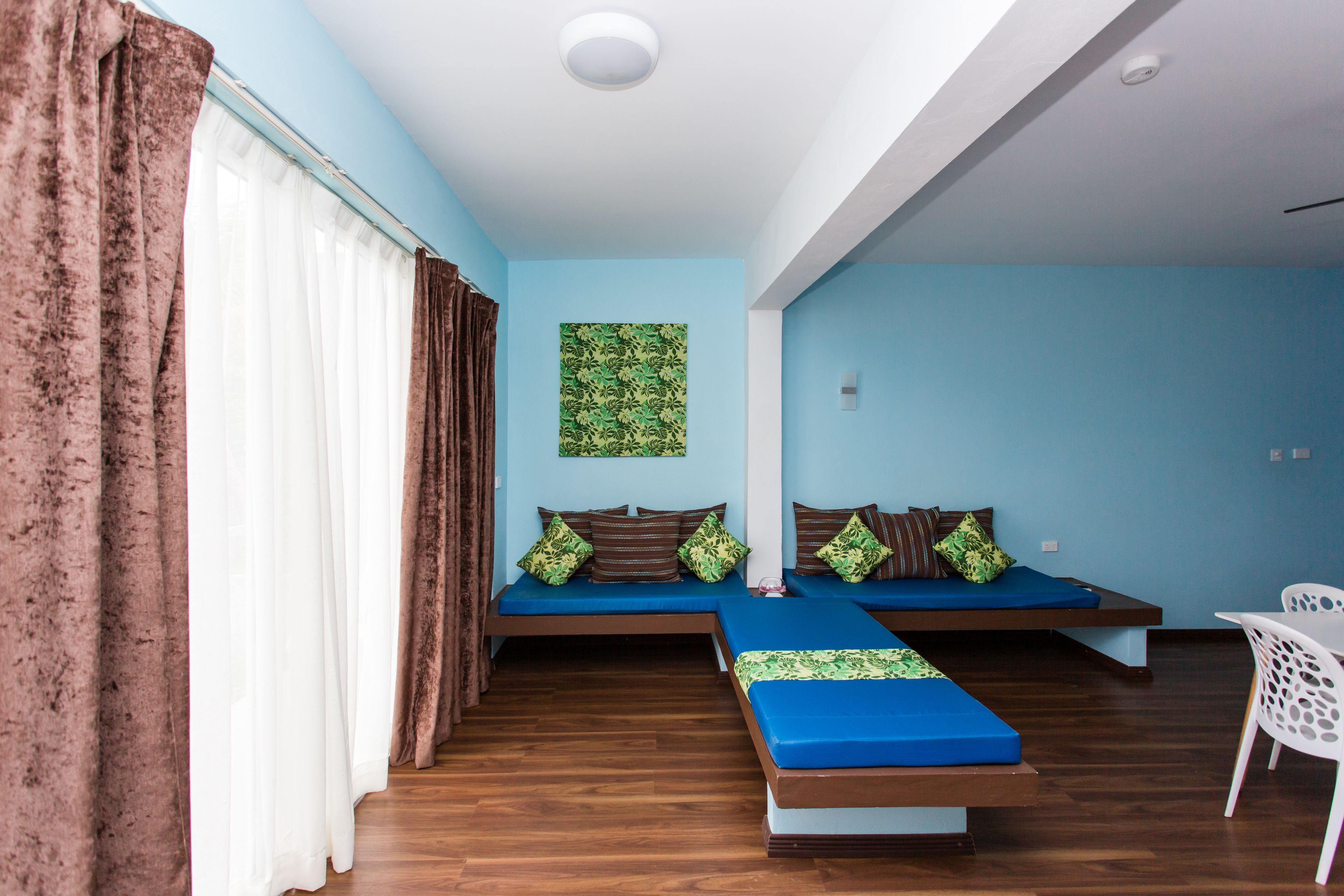 Una habitación con pared azul, cama con ropa de cama azul, una silla blanca y una ventana con cortinas marrones.