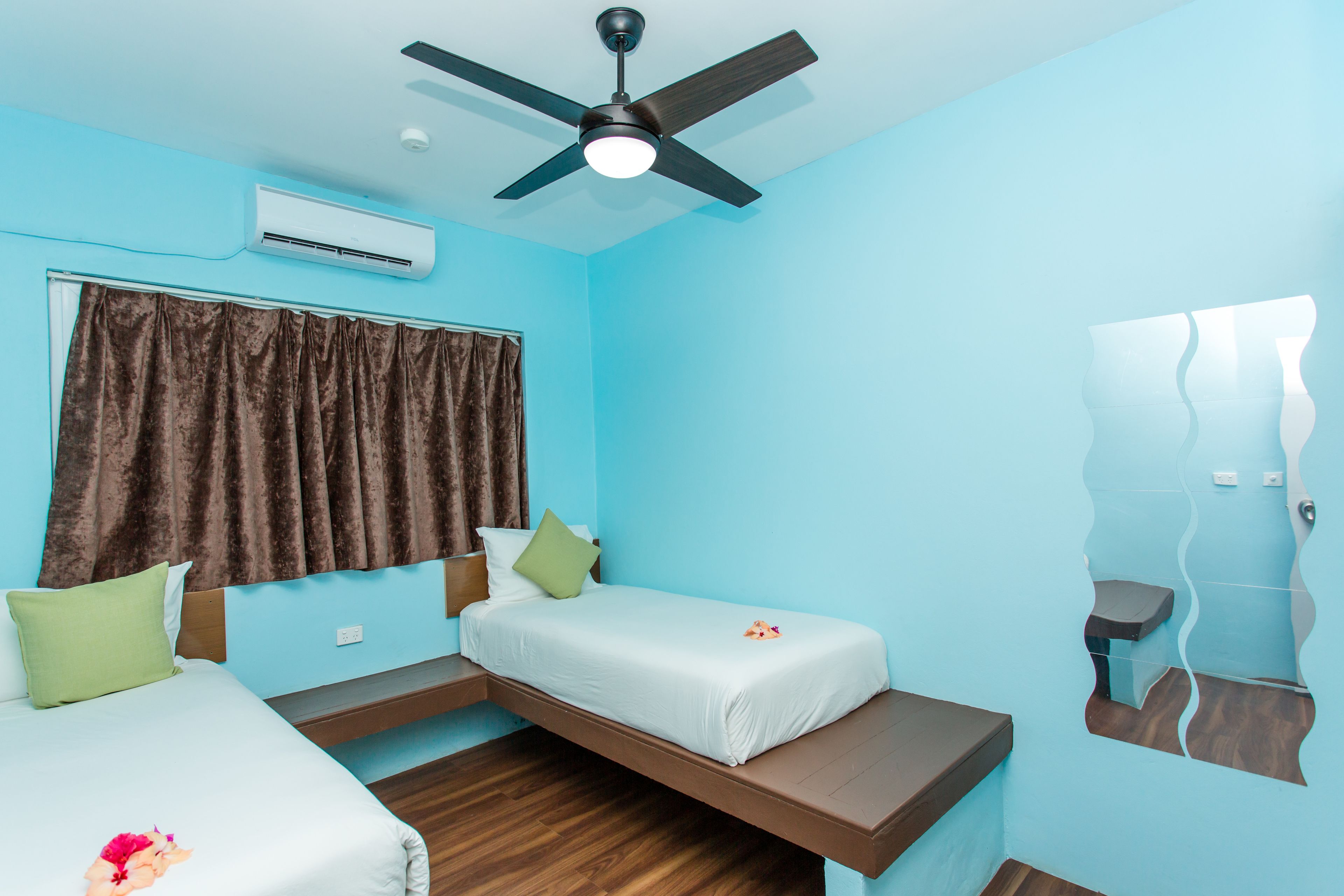 Una habitación con dos camas, ventilador de techo, aire acondicionado, una ventana con cortinas y un espejo en la pared.