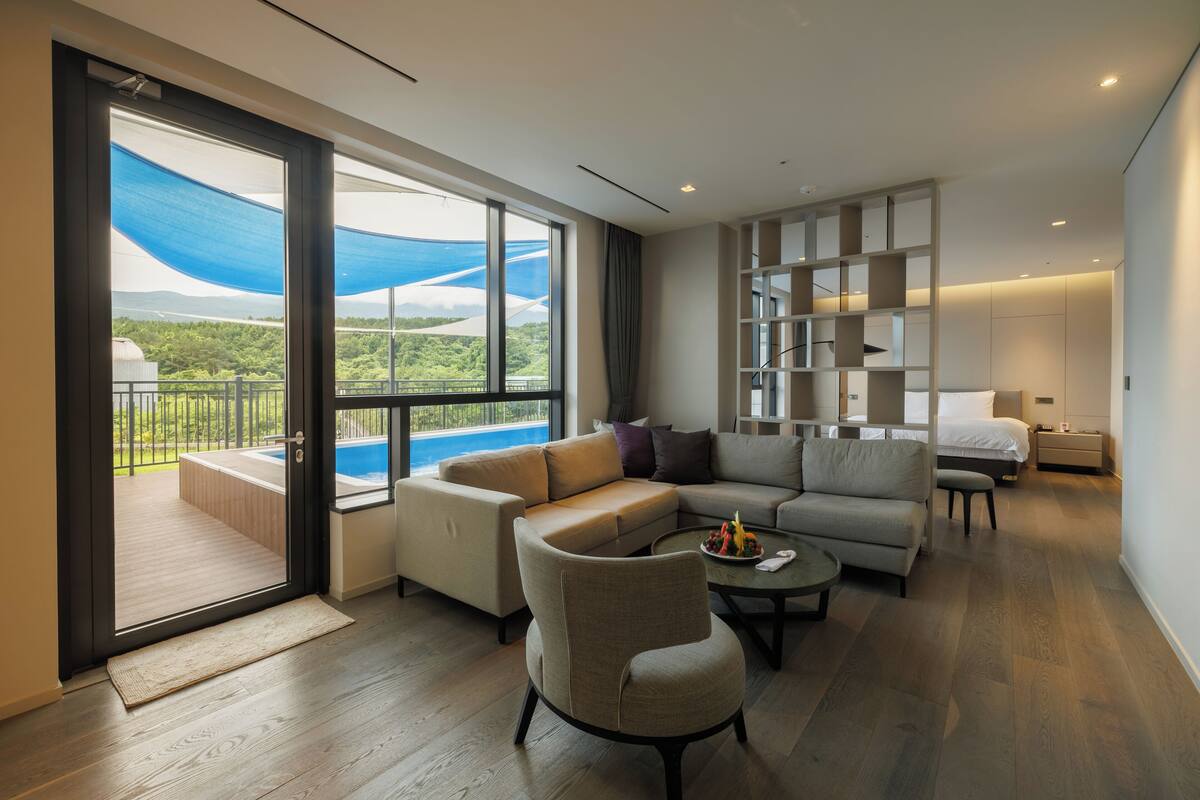 A modern living room with a large sliding glass door leading to a balcony, a sectional sofa, a small round table, and a built-in shelving unit.