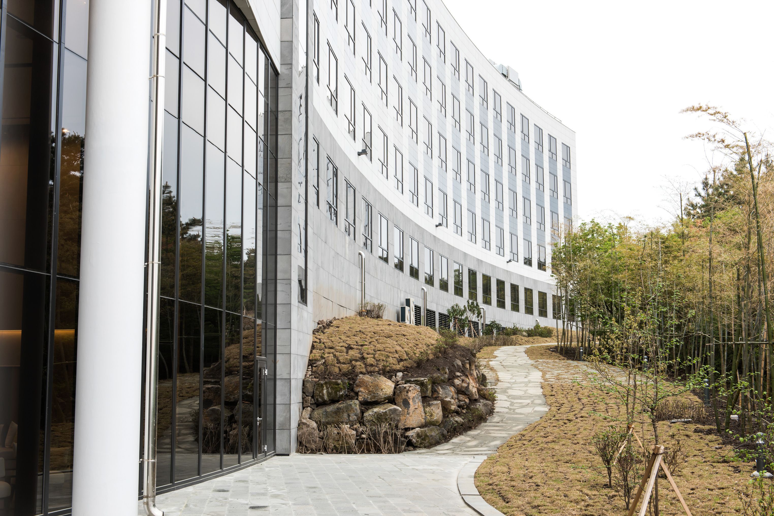 A modern building with a curved facade, large windows, and a stone pathway leading to a landscaped area with rocks and sparse vegetation.