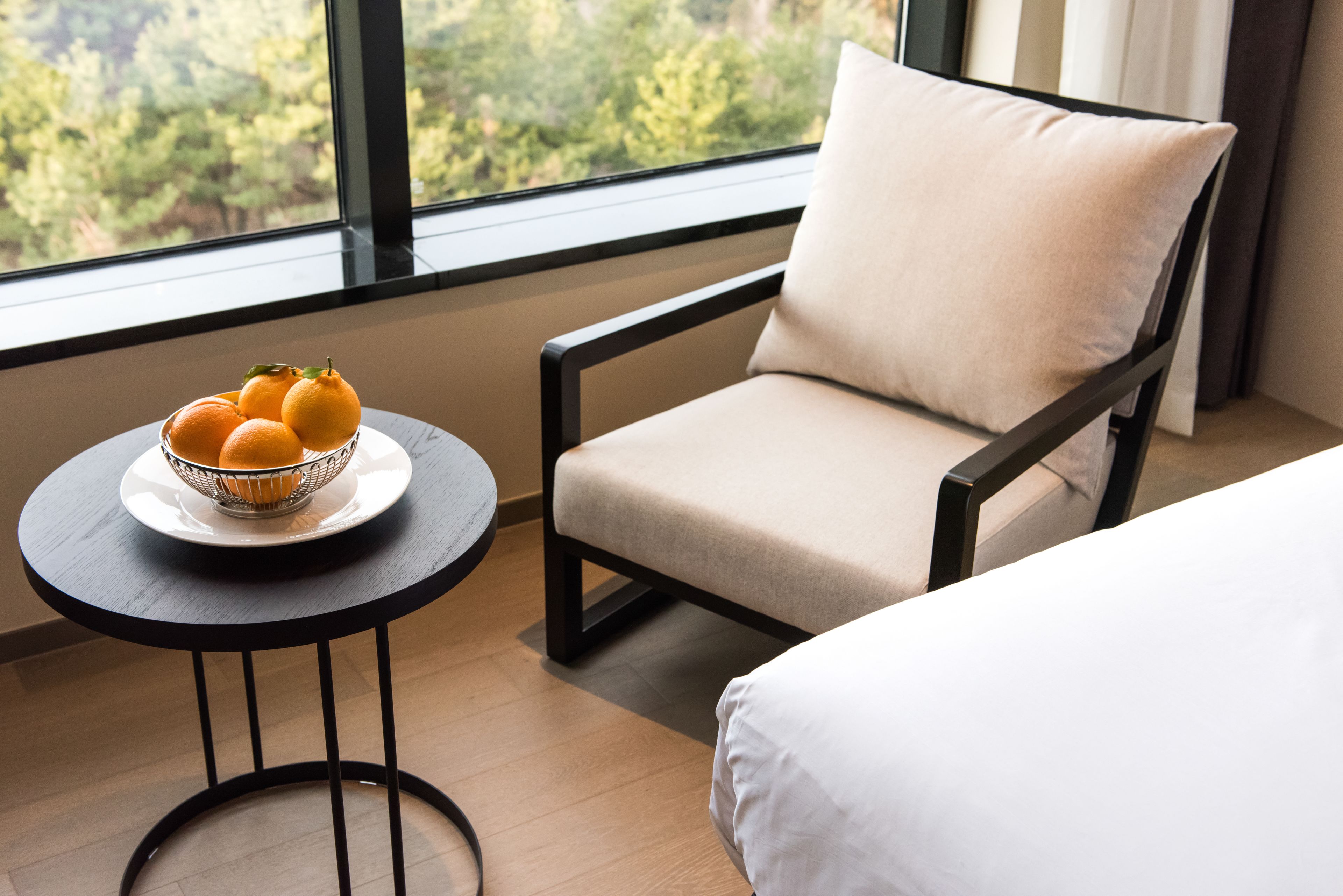 Une chambre d’hôtel avec vue sur les arbres, visible à travers une grande fenêtre, une table ronde avec un saladier rempli d’oranges et une chaise avec un coussin.