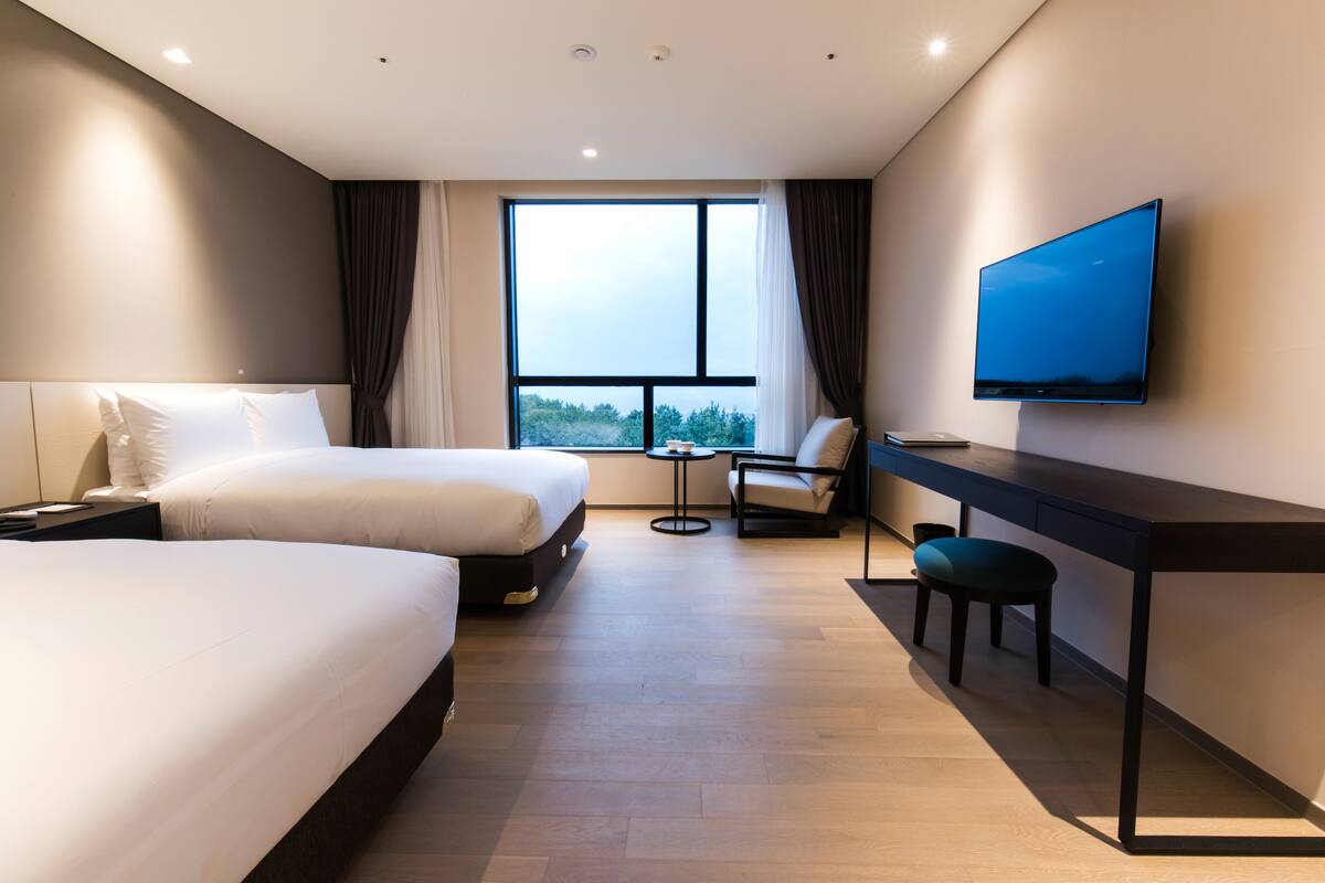 A modern hotel room with two beds, a desk, a chair, a TV, and a large window offering a view of the outdoors.