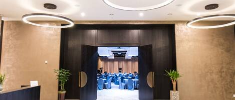 Un moderno vestíbulo de hotel con mostrador de recepción, plantas en macetas y vistas a un salón de banquetes con mesas y sillas redondas.