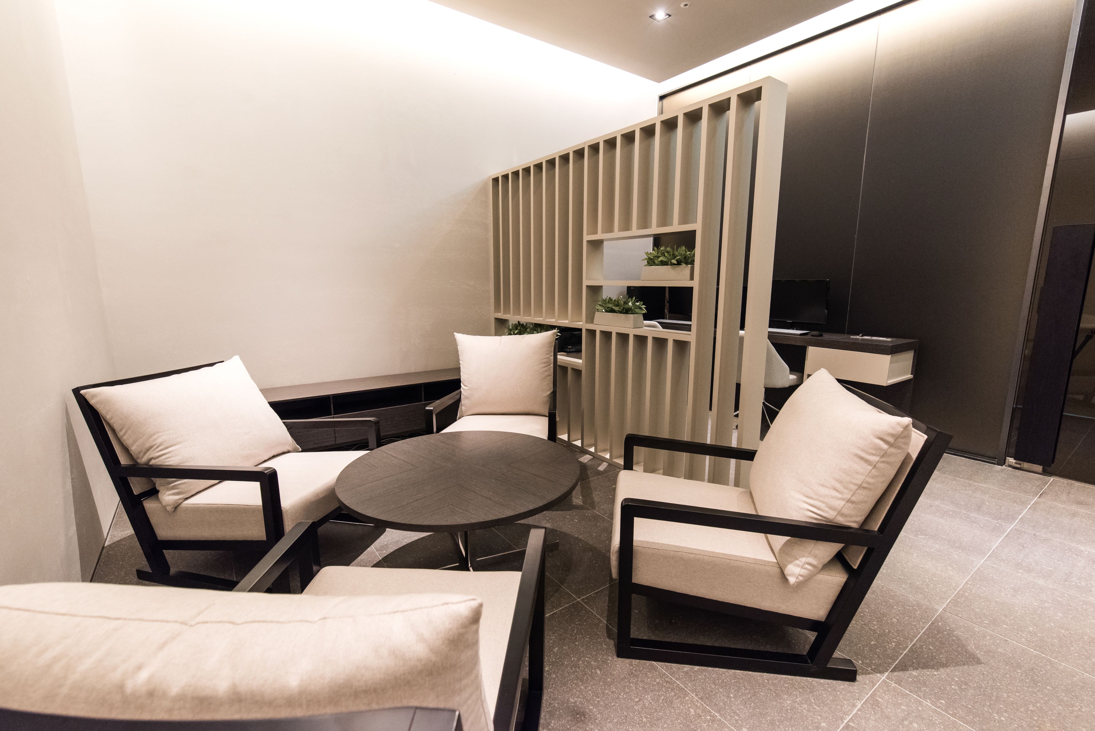 A modern living room with a round table, two armchairs, and a sofa. There is a built-in TV shelf and a small plant on the wall.