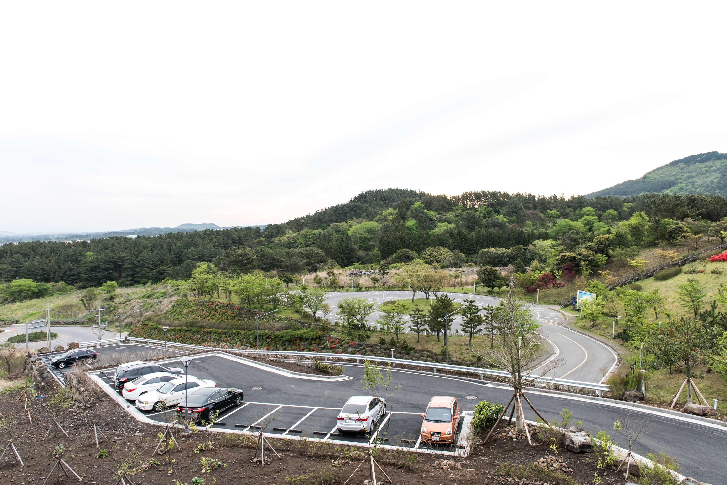A parking lot with several cars, a winding road, and a hilly landscape with trees.