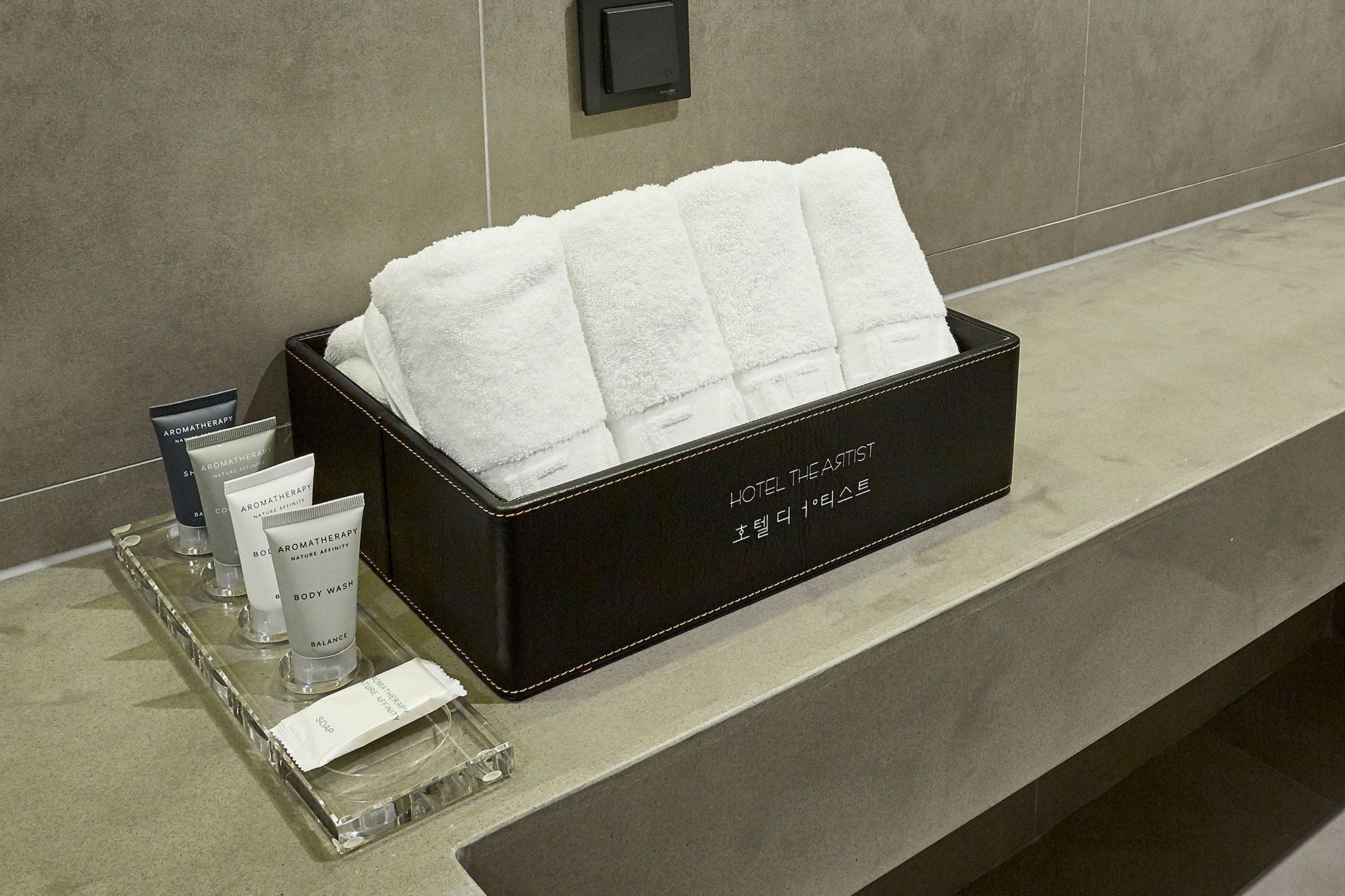 A bathroom counter with a black box containing white towels, a body wash, and two tubes of soap.