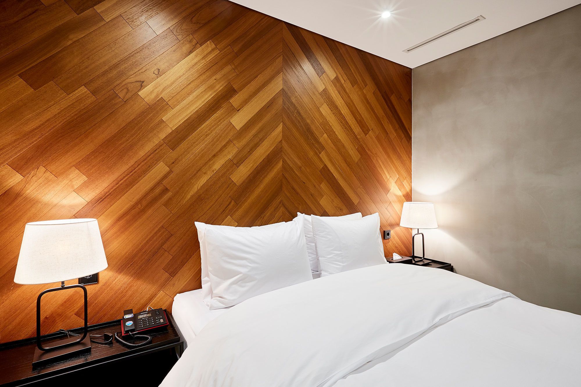 A hotel room with a wooden herringbone wall, a bed with white linens, two bedside lamps, and a telephone on a nightstand.