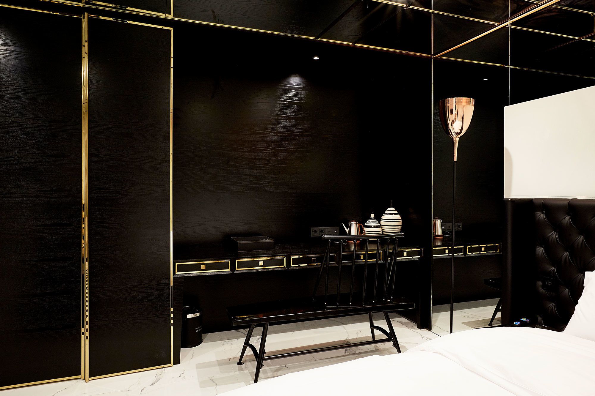 Een moderne slaapkamer met een zwart en goud kleurenschema, een bed met een wit hoofdeinde, een bureau met een lamp en een grote spiegel.