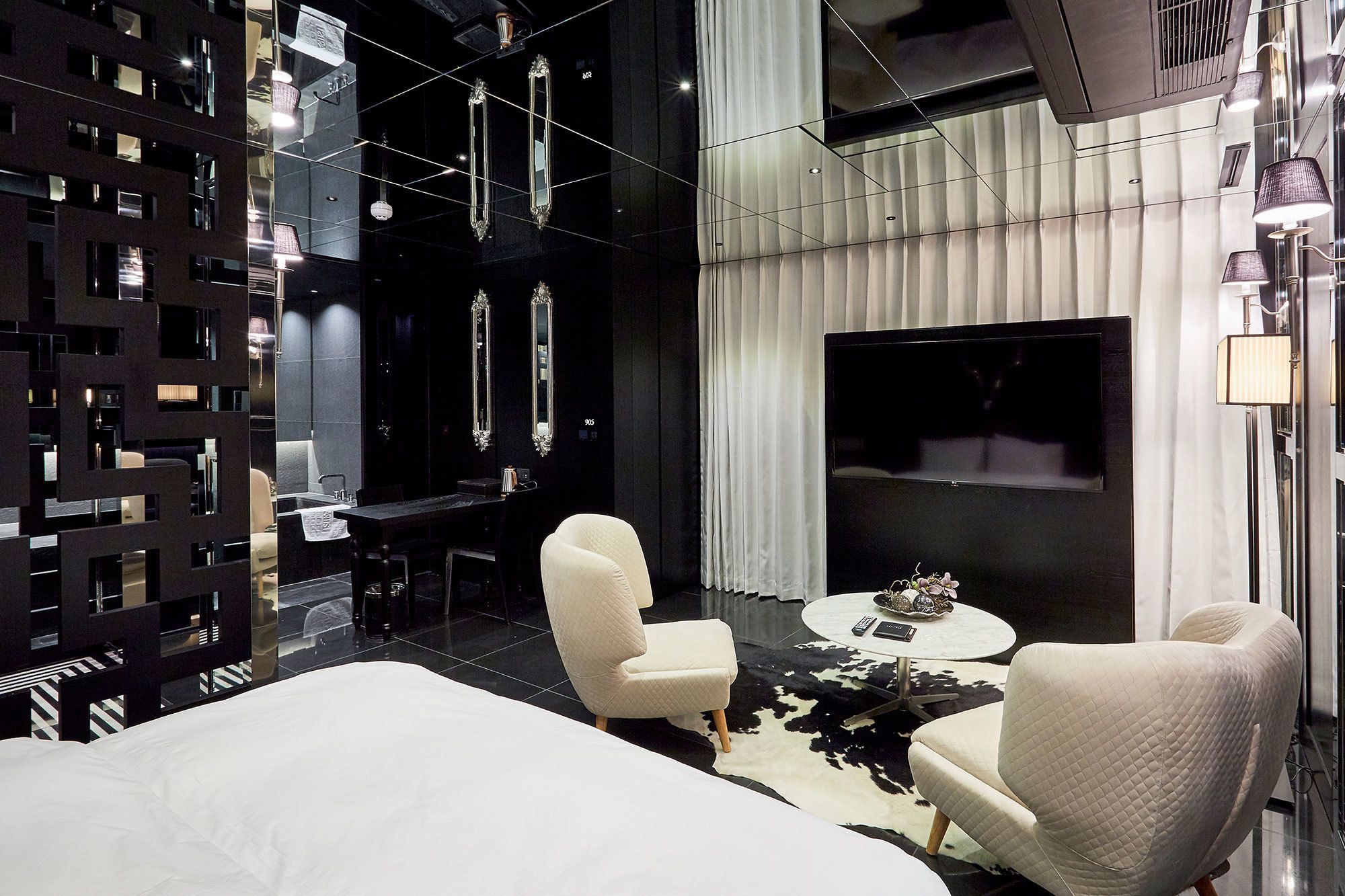 Een moderne hotelkamer met een groot bed, twee witte fauteuils, een ronde salontafel en een grote flatscreen-tv.