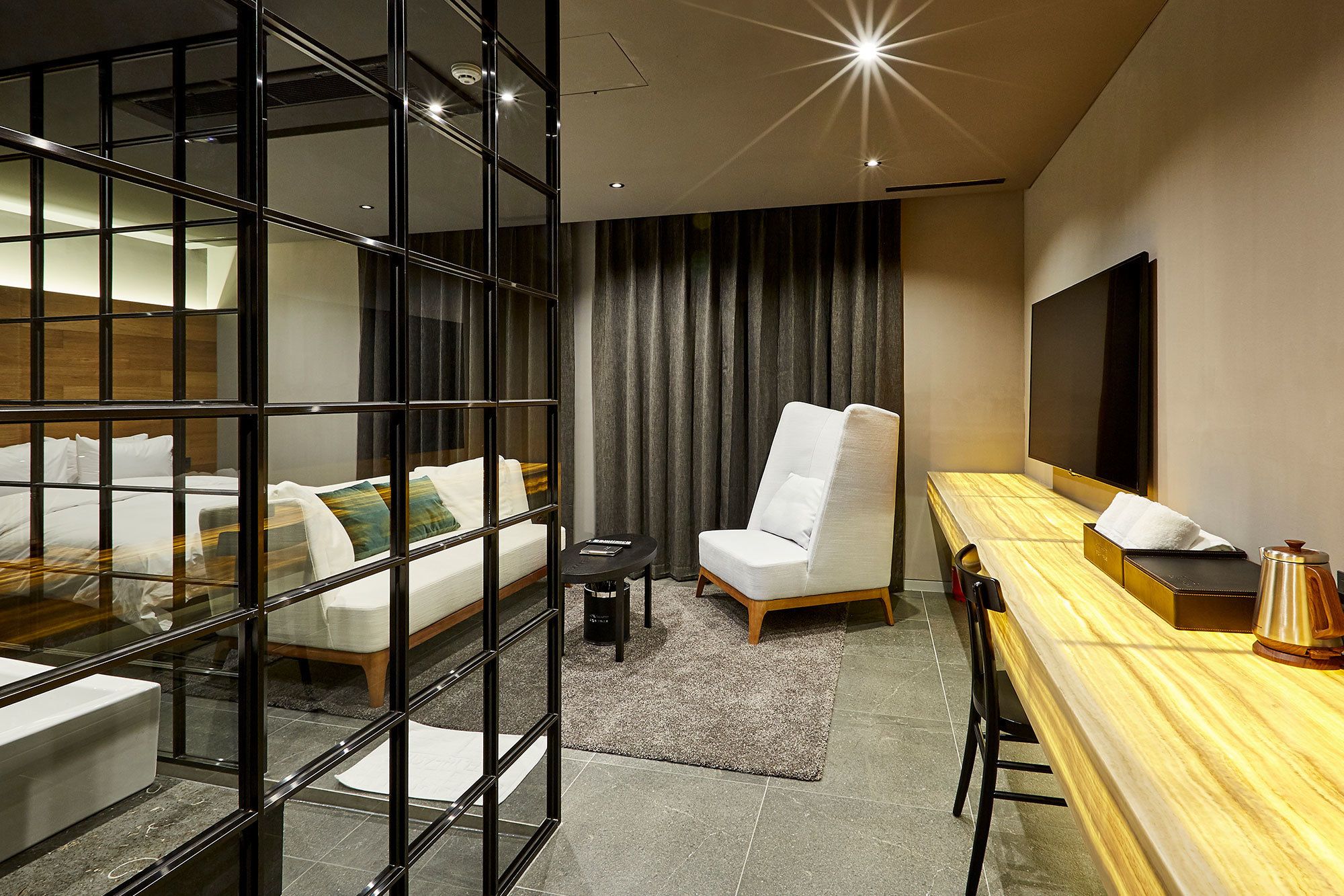 A modern hotel room with a glass partition separating the bed area from the seating area, a large flat-screen TV, a wooden table with a coffee pot, and a comfortable chair.