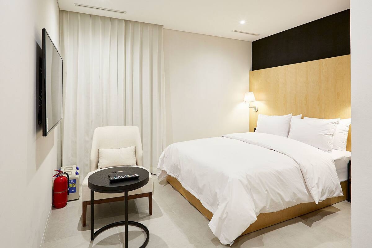 A hotel room with a bed, a chair, a small table, a fire extinguisher, and a wall-mounted TV.