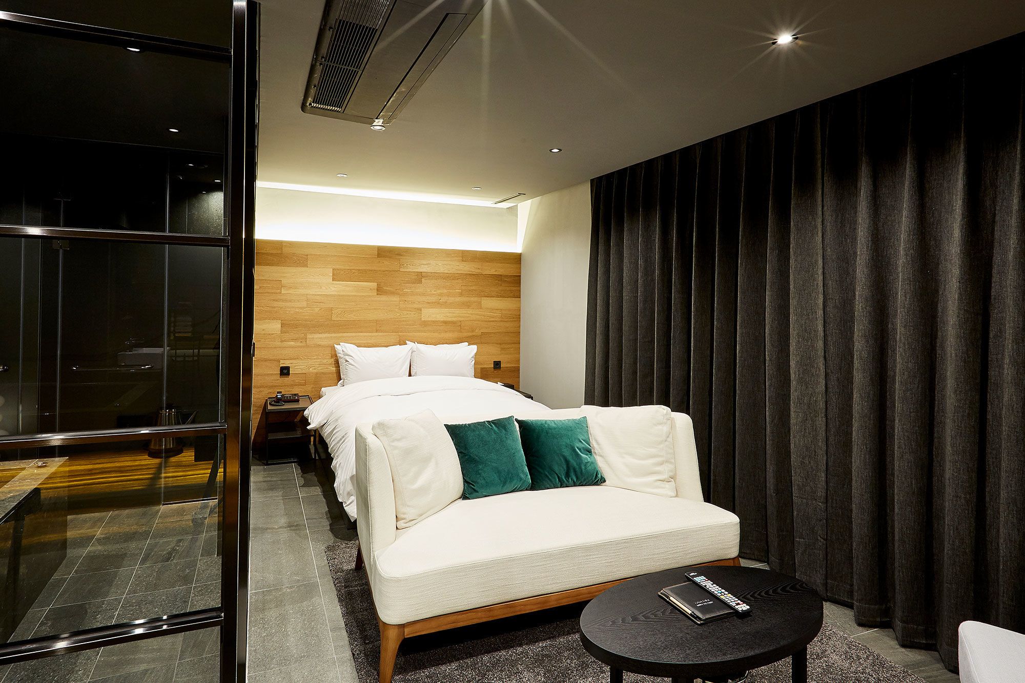 Habitación de hotel moderna con cabecera de madera, sofá blanco con cojines verdes, una pequeña mesa redonda y un separador de vidrio.