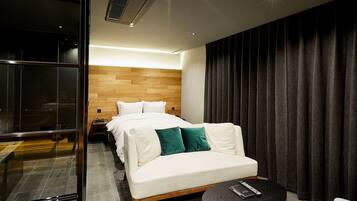 Habitación de hotel moderna con cabecera de madera, sofá blanco con cojines verdes, una pequeña mesa redonda y un separador de vidrio.
