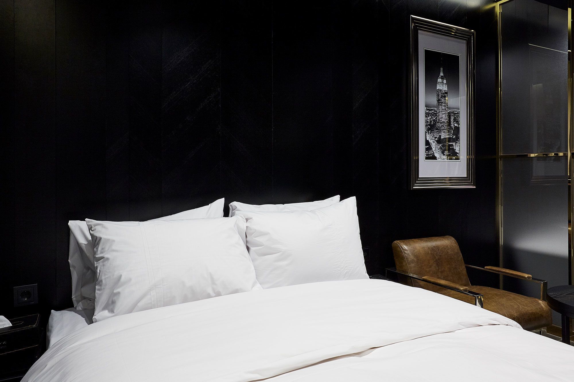 A neatly made bed with white linens in a dark room with a framed picture of a city skyline on the wall.