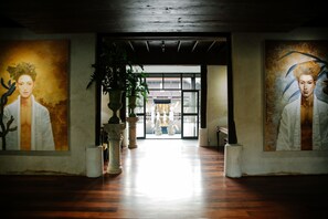 A hallway with wooden flooring, large paintings on the walls, and a potted plant in the center.