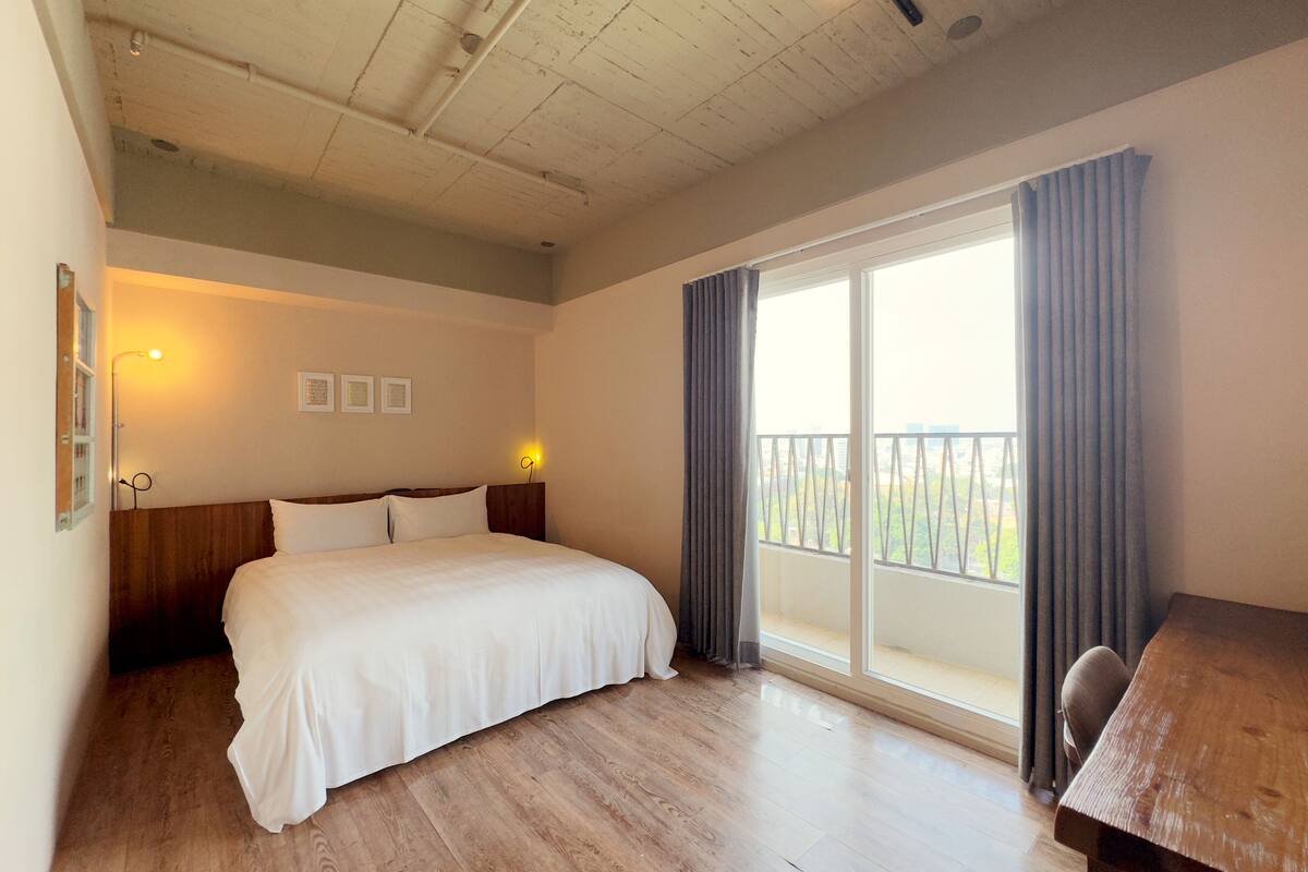 Ein Hotelzimmer mit Holzboden, einem Bett mit weißer Bettwäsche, einem Holzkopfende, einem Schreibtisch und einem Balkon mit Vorhängen.