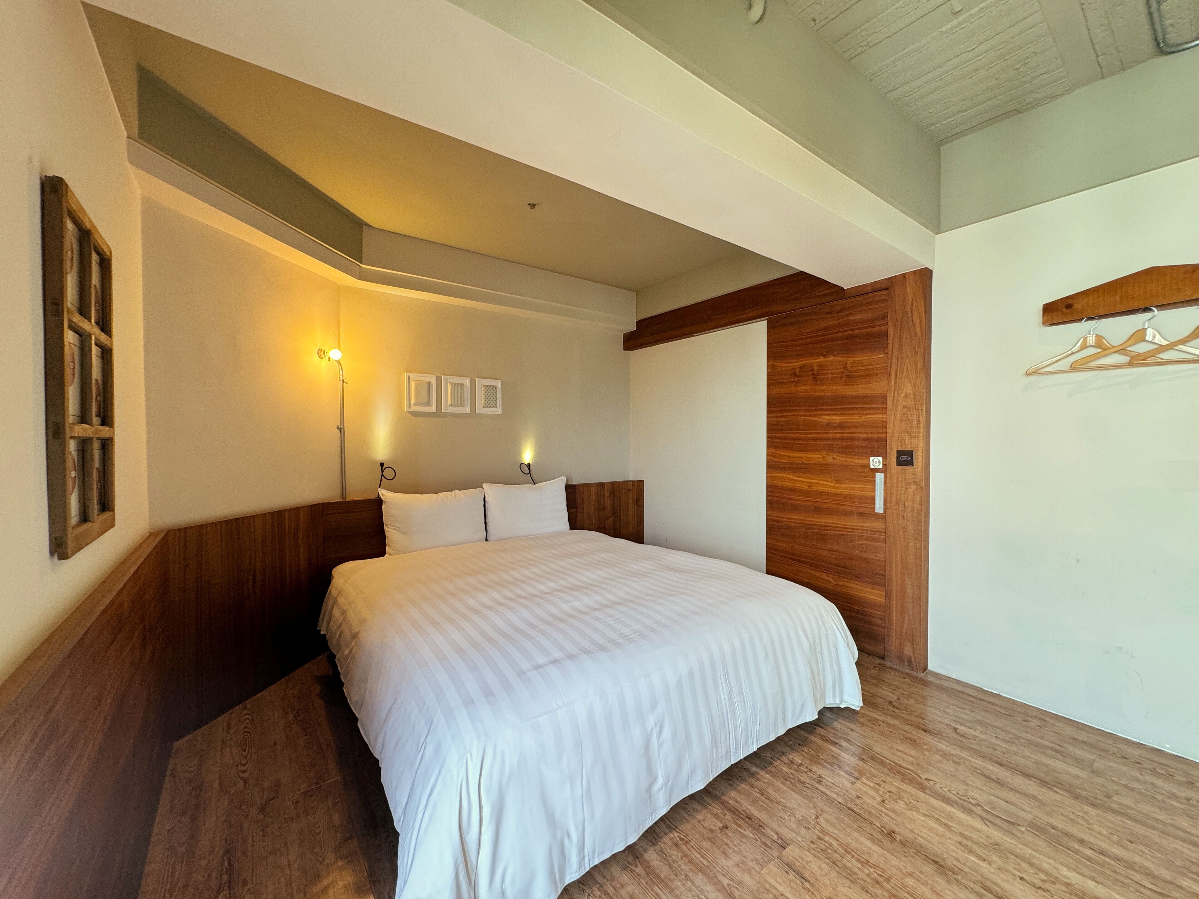Un dormitorio con cabecera de madera, una cama grande con ropa de cama blanca, una lámpara fijada a la pared y una puerta de madera.