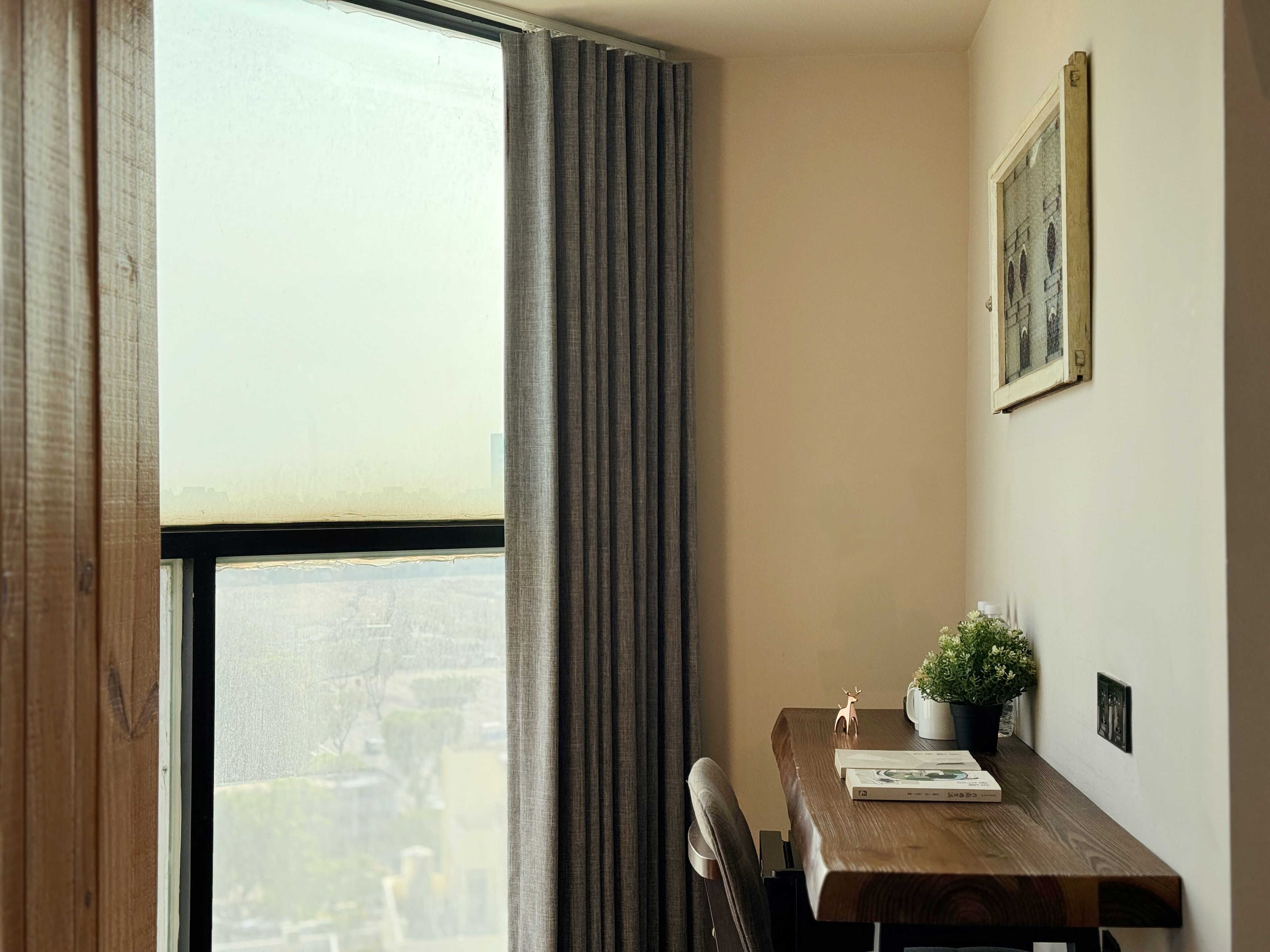 A corner room with a wooden table, a potted plant, a framed picture on the wall, and a window with curtains.