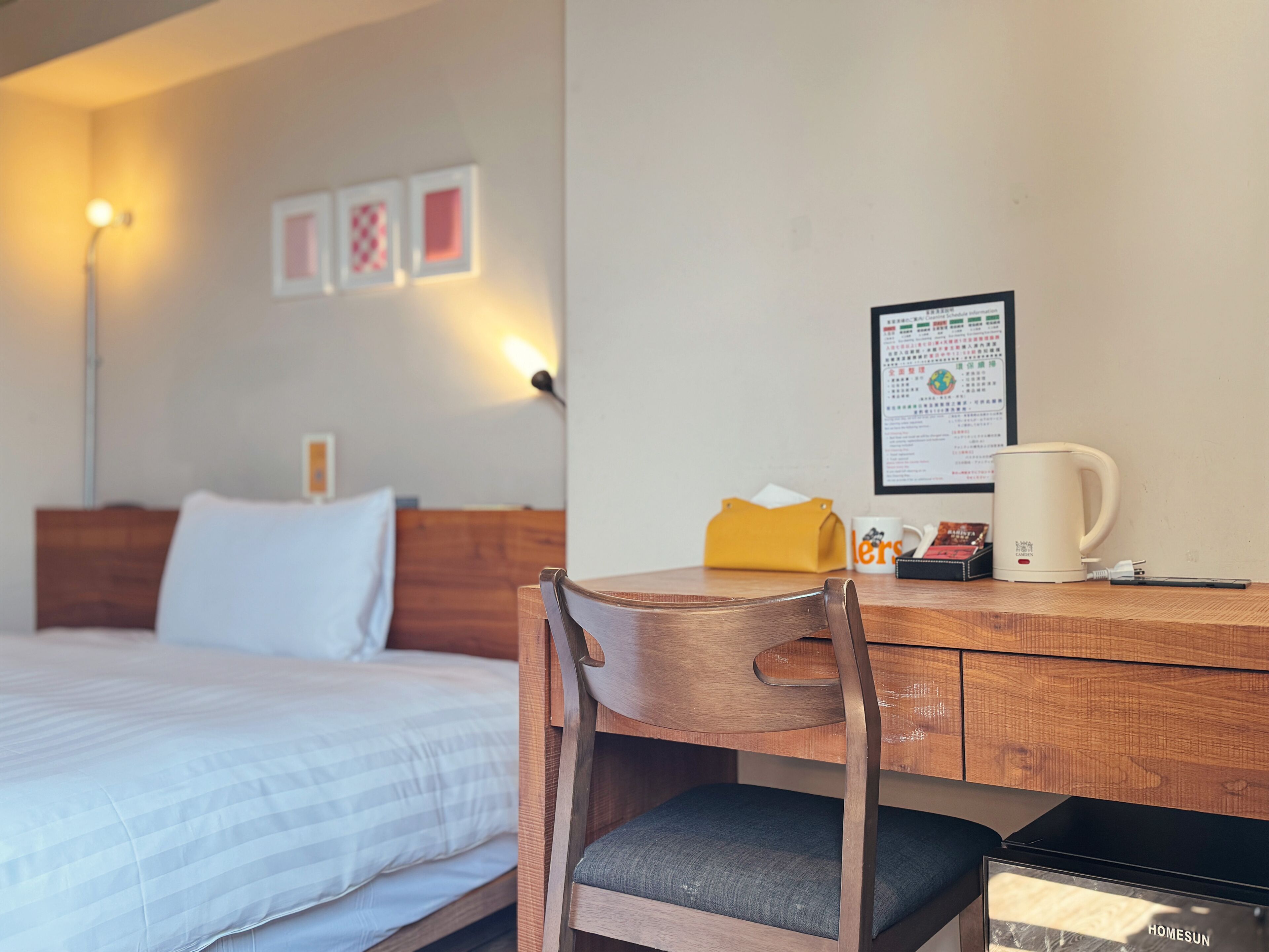 A hotel room with a bed, a wooden desk, a chair, a coffee maker, and a framed menu on the wall.