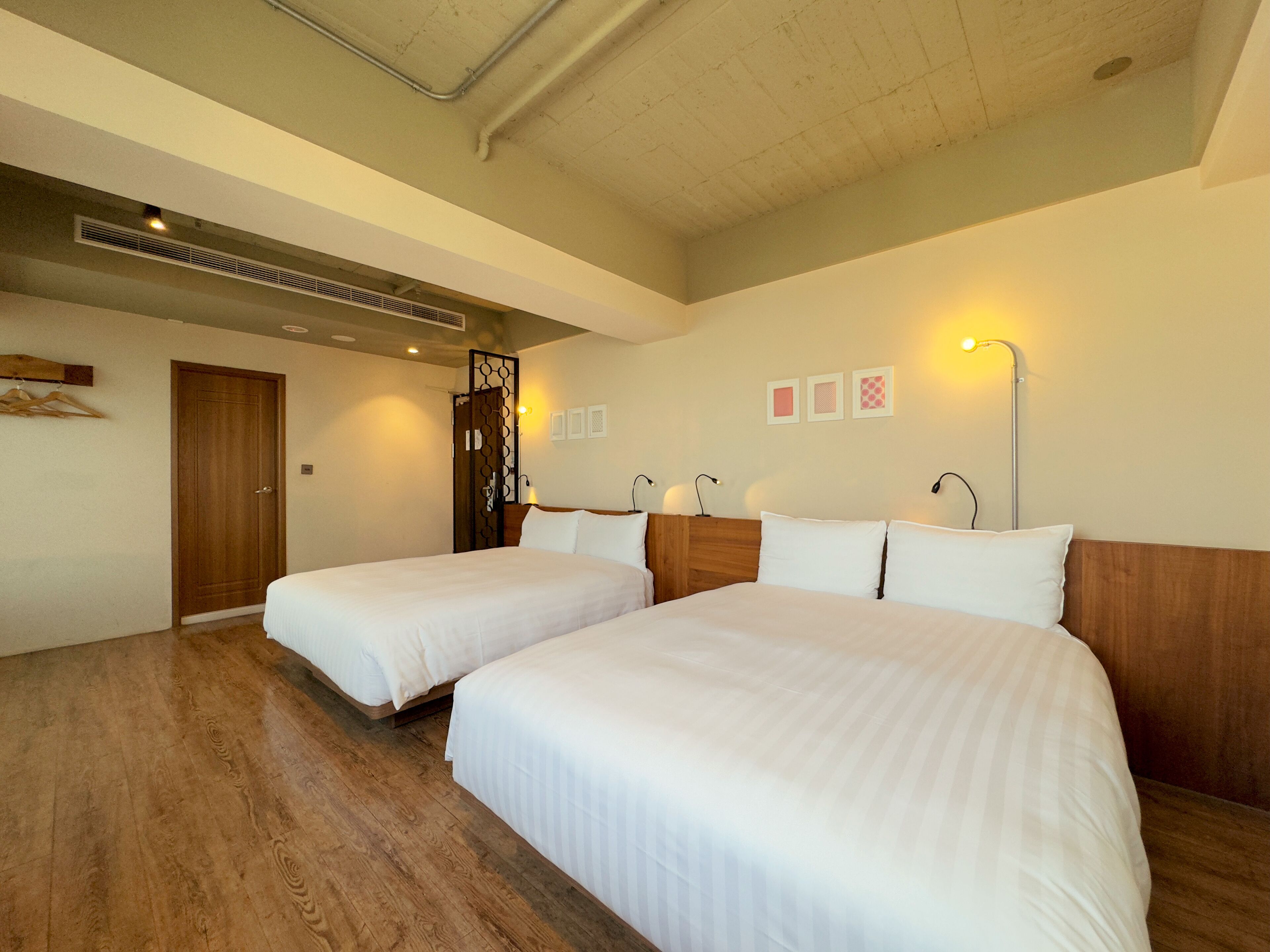 Quarto de hotel com duas camas de solteiro, piso de madeira, porta de madeira e teto com tubulação aparente.