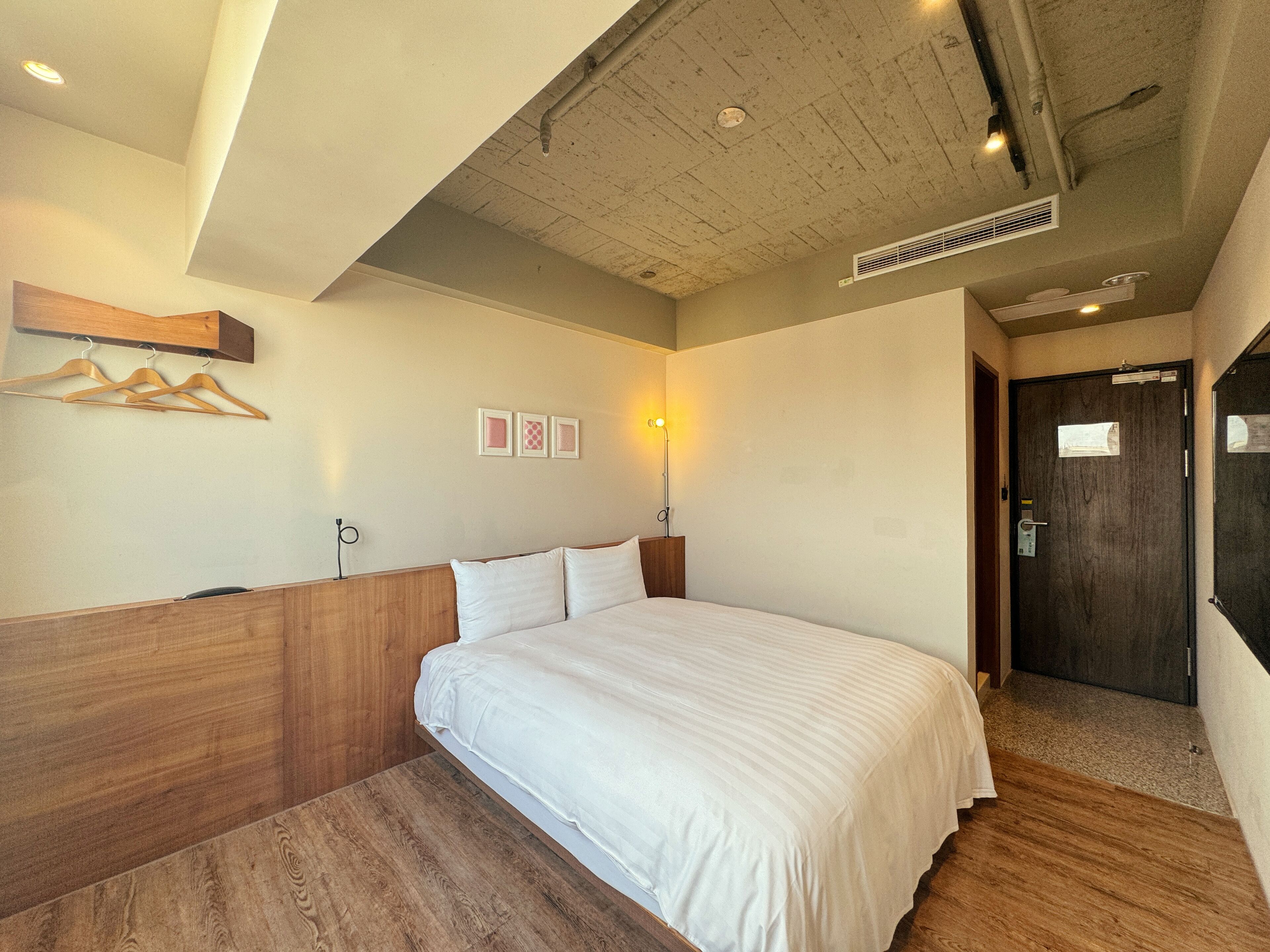 A hotel room with a wooden headboard, a large bed with white bedding, a wall-mounted light, and a door with a mirror and a sign.