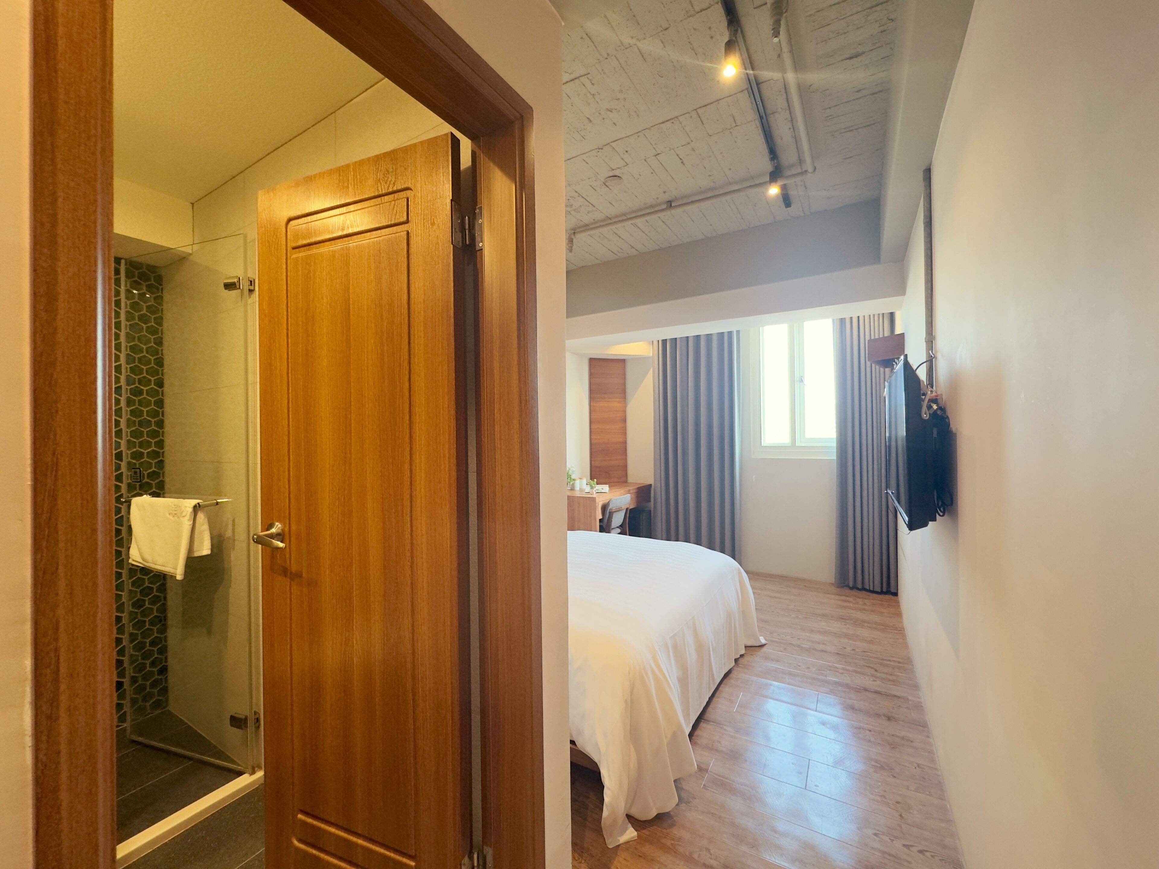 A hotel room with a wooden door, a shower with a glass door, a bed with white bedding, a desk, and a window with curtains.