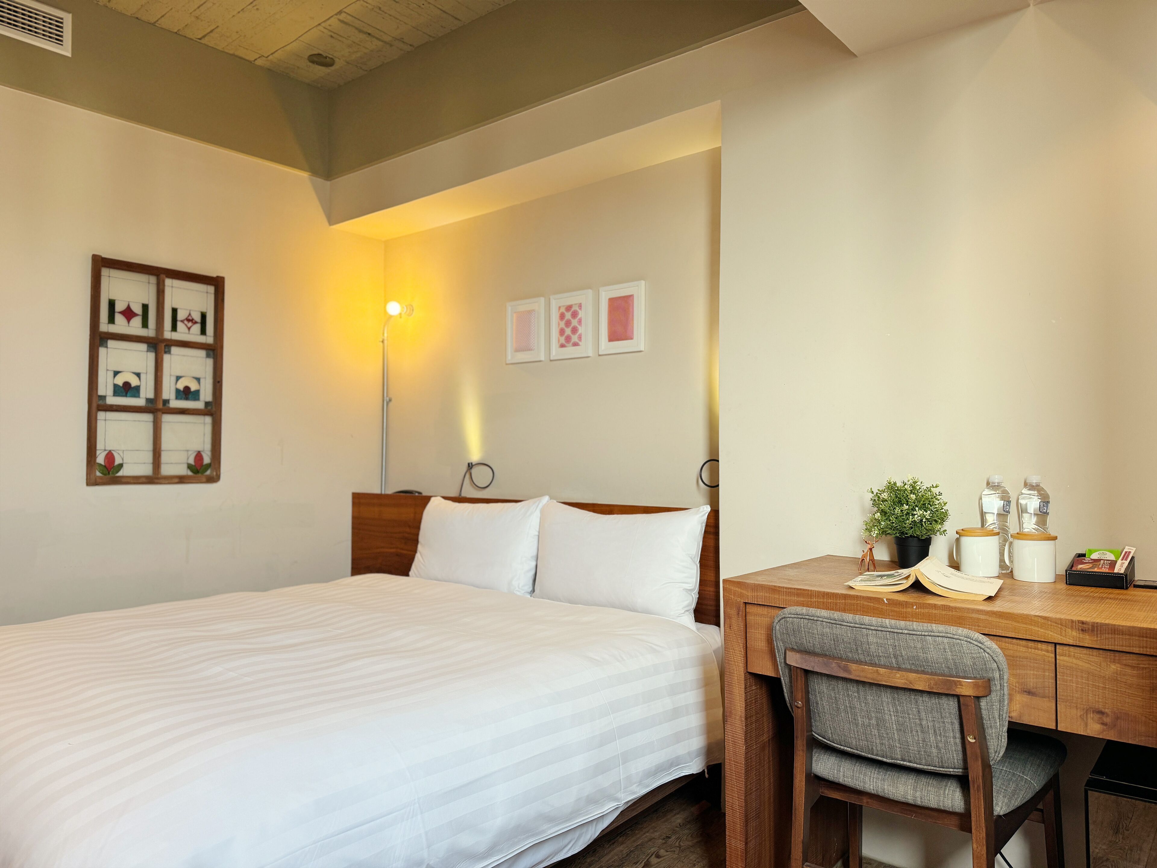 A neatly arranged bedroom with a bed, bedside table, chair, and a desk with a plant and water bottles.