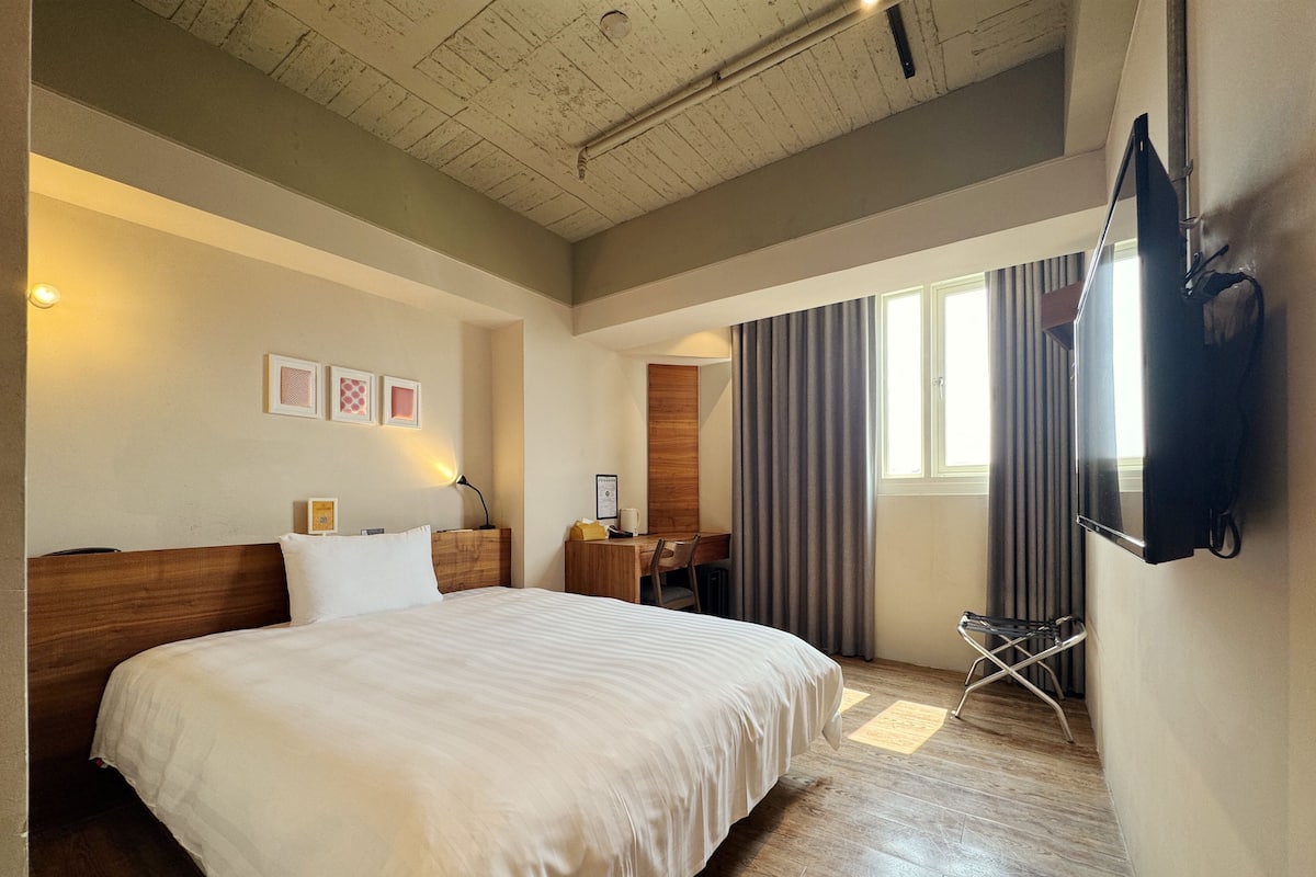 Ein Hotelzimmer mit einem großen Bett, einem Schreibtisch, einem Stuhl, einem Fernseher und einem Fenster mit Vorhängen.