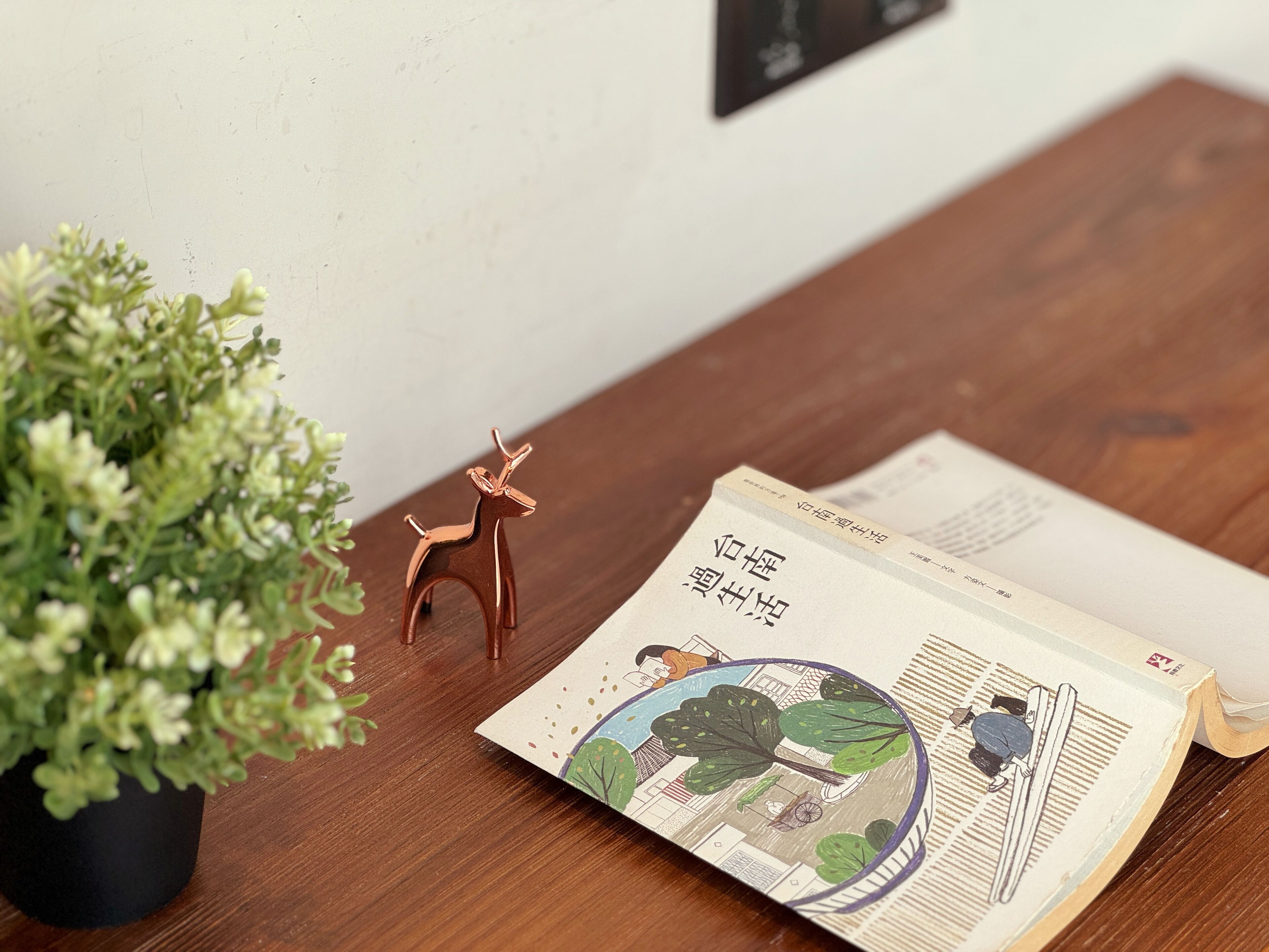 A wooden table with a potted plant, a small deer figurine, and an open book with illustrations.