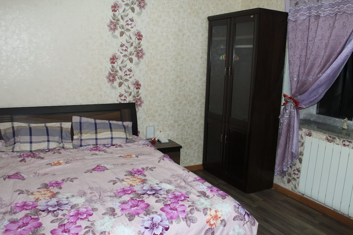 A bedroom with a floral bedspread, a dark wooden wardrobe, and a window with a purple curtain.