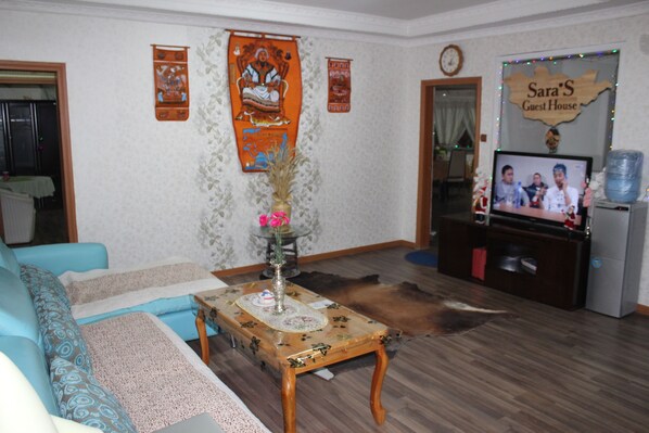 A living room with a sofa, a coffee table with a vase, a television, and a sign reading "Sara's Guest House".