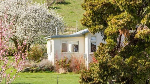 A small, single-story house with a sloped roof, surrounded by lush greenery and flowering trees.