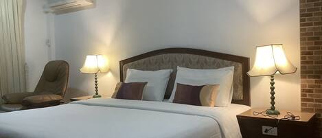 Quarto de hotel com uma cama grande, duas luminárias de cabeceira, uma cadeira e um aparelho de ar condicionado.