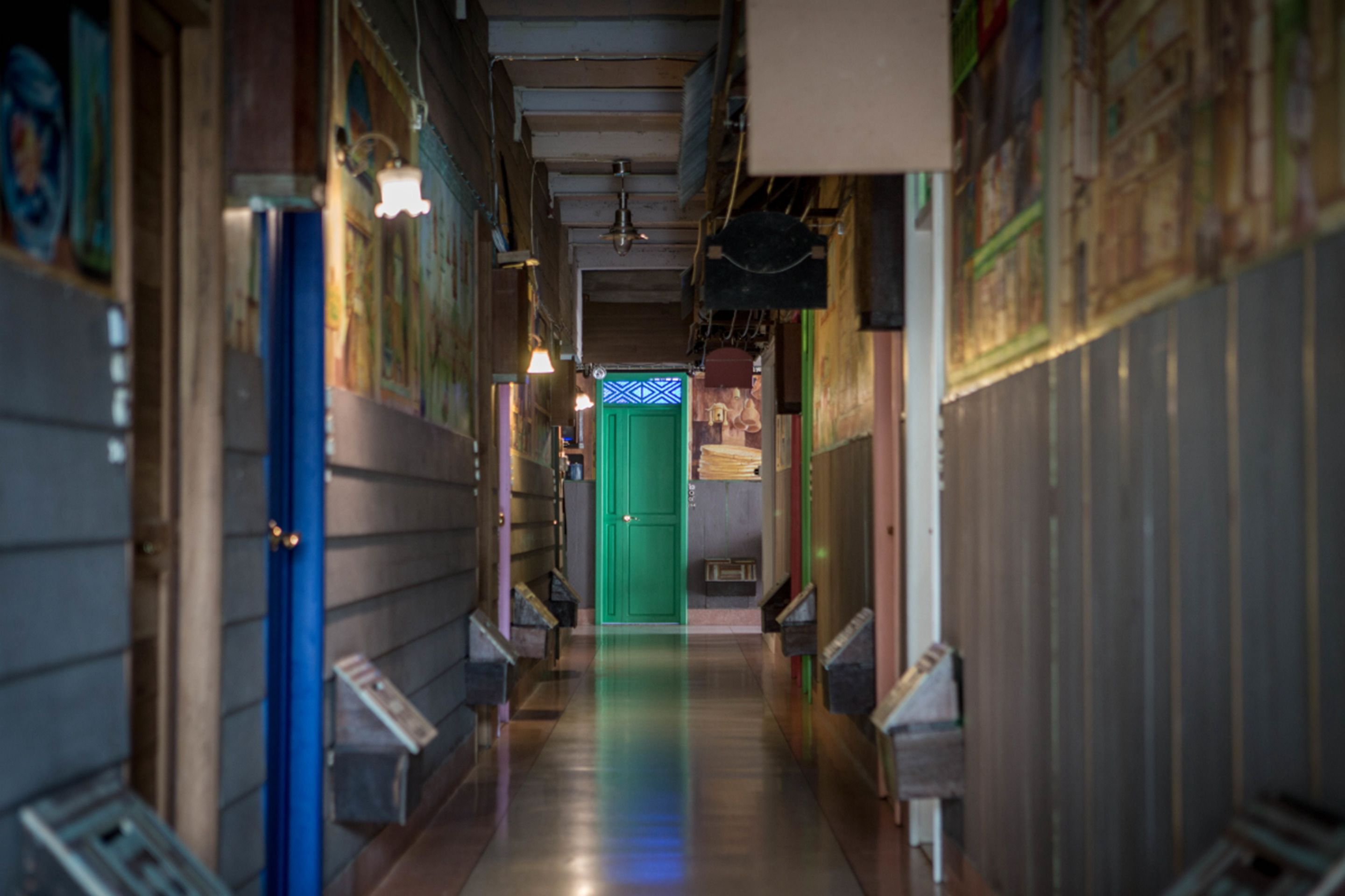 Un pasillo estrecho con una puerta verde al final, flanqueado por paredes con diversas decoraciones y una barandilla de metal a la izquierda.