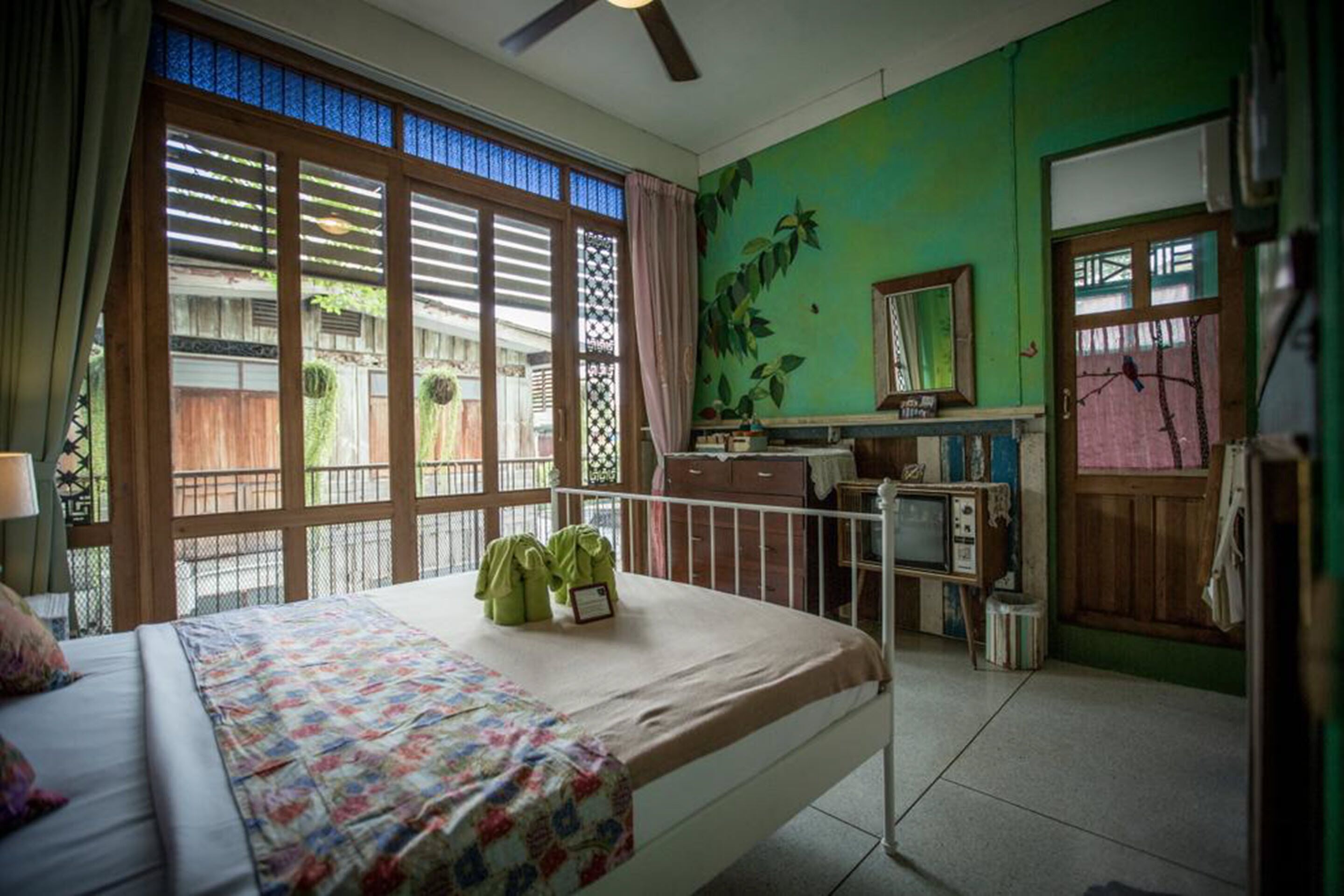 A room with a bed, a desk with a television, and a green wall with a plant mural.