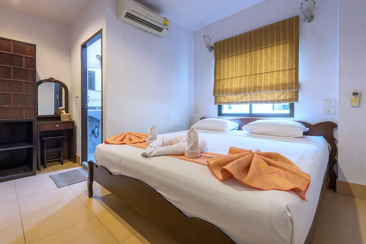 A hotel room with a bed, a wooden nightstand, a mirror, and a window with a blind.