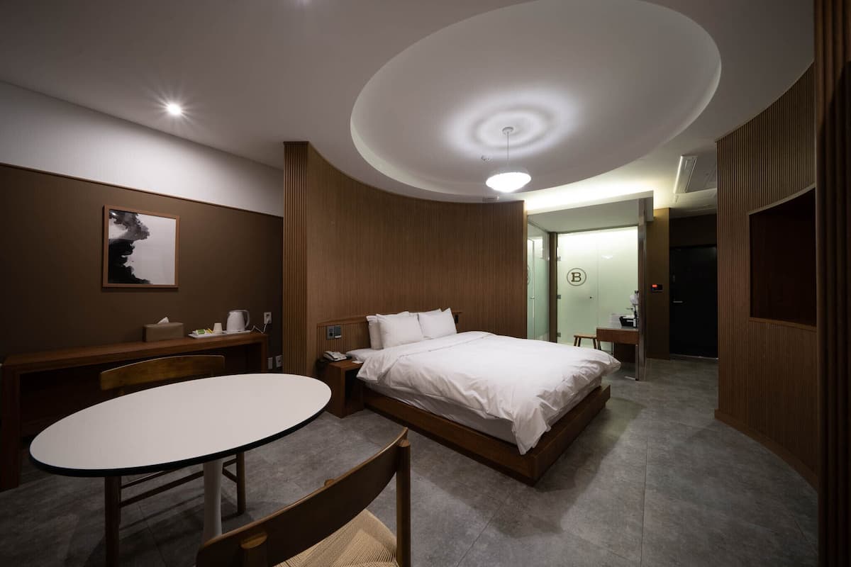 A modern hotel room with a large bed, a desk, and a chair. There is a painting on the wall and a door with a 'B' sign.