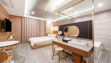 Una habitación de hotel moderna con cama, escritorio, silla, un espejo grande y baño con lavabo e inodoro.