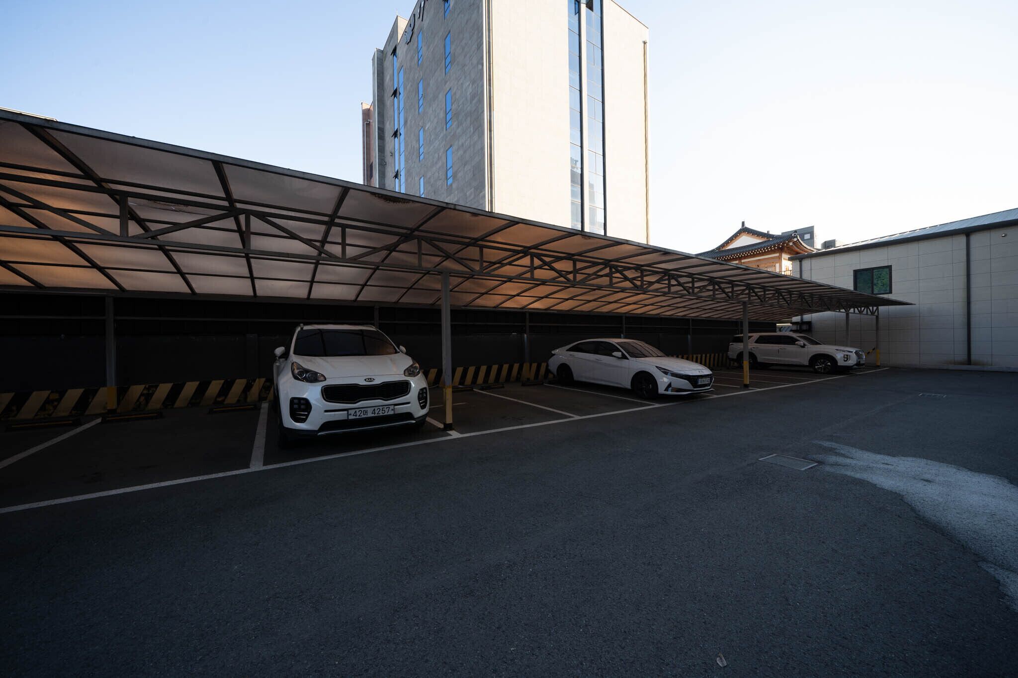 Uma garagem com área coberta, dois carros brancos estacionados e um edifício com grandes janelas ao fundo.