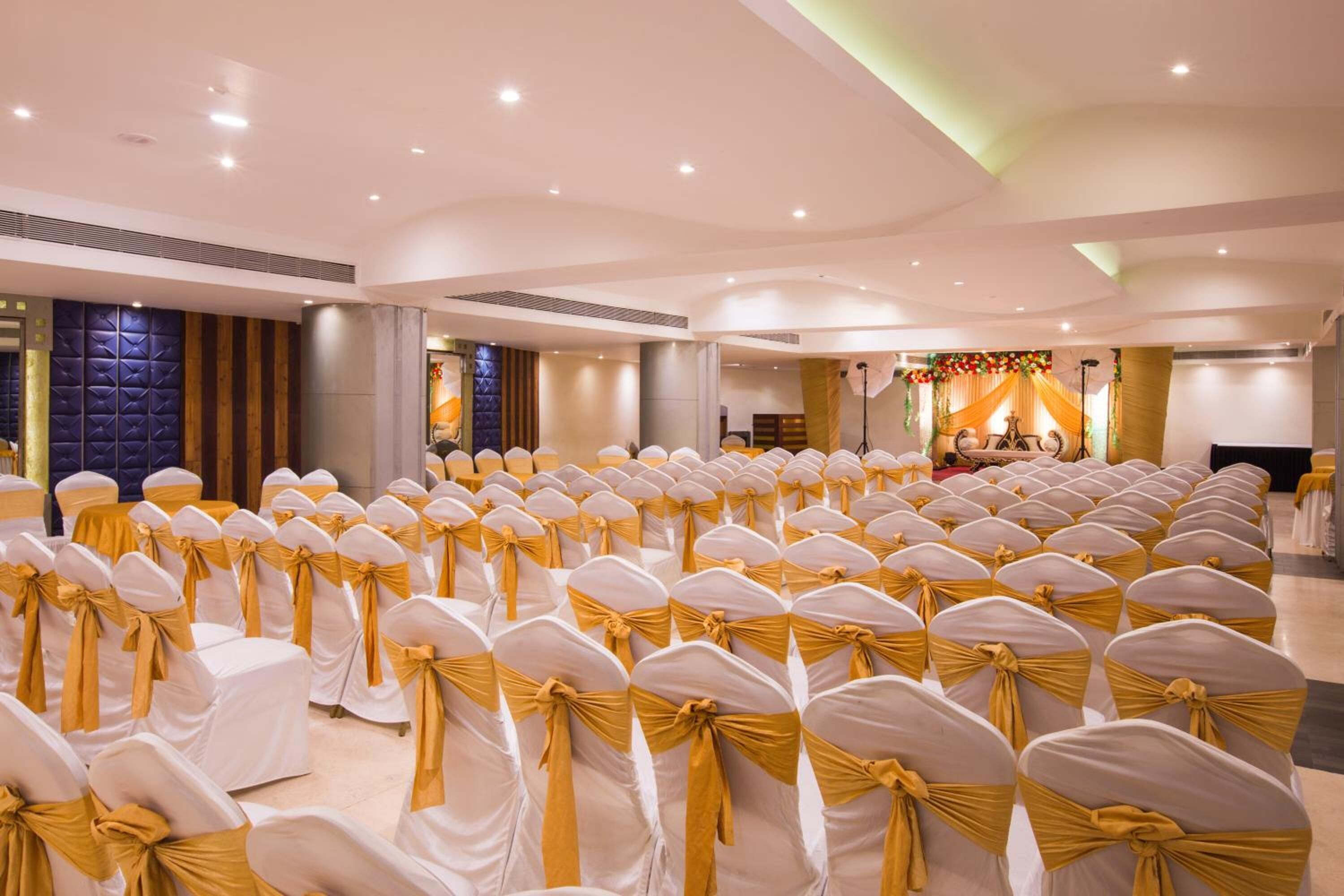 Ein großer Saal mit Reihen aus weißen Stühlen mit goldenen Bändern, einer Bühne mit Bühnenbild und einer Decke mit eingelassener Beleuchtung.