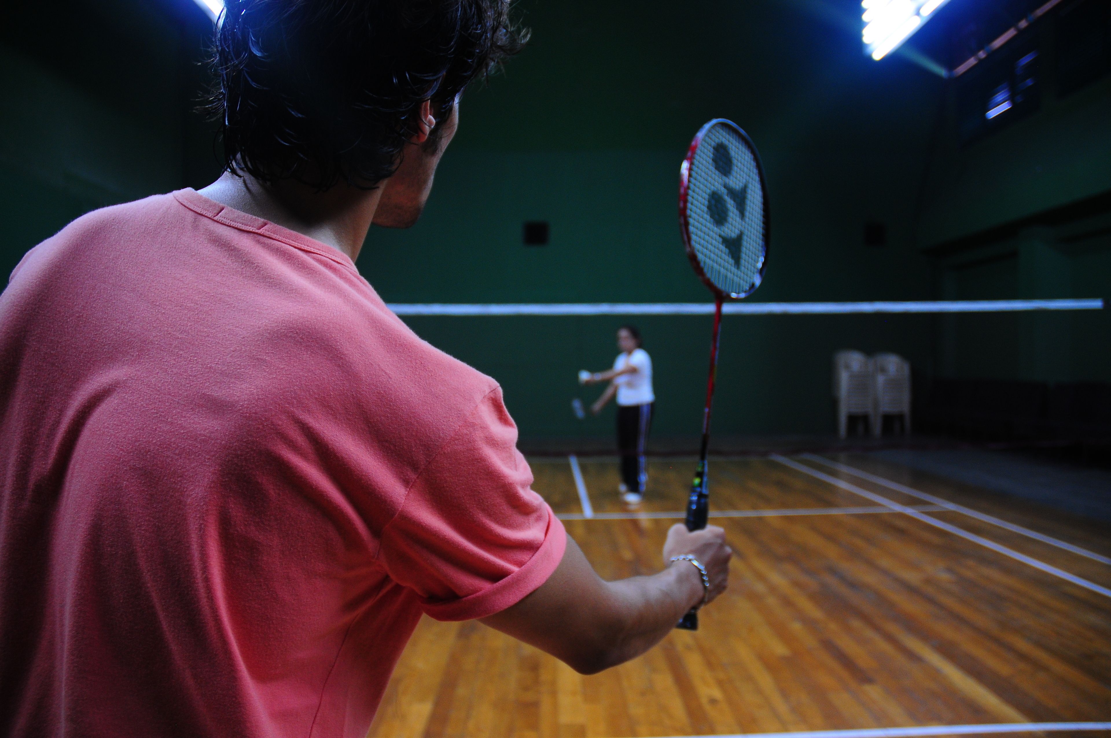 En person i en rosa tröja som håller i en badmintonracket i en gymnasal med trägolv och ett nät.