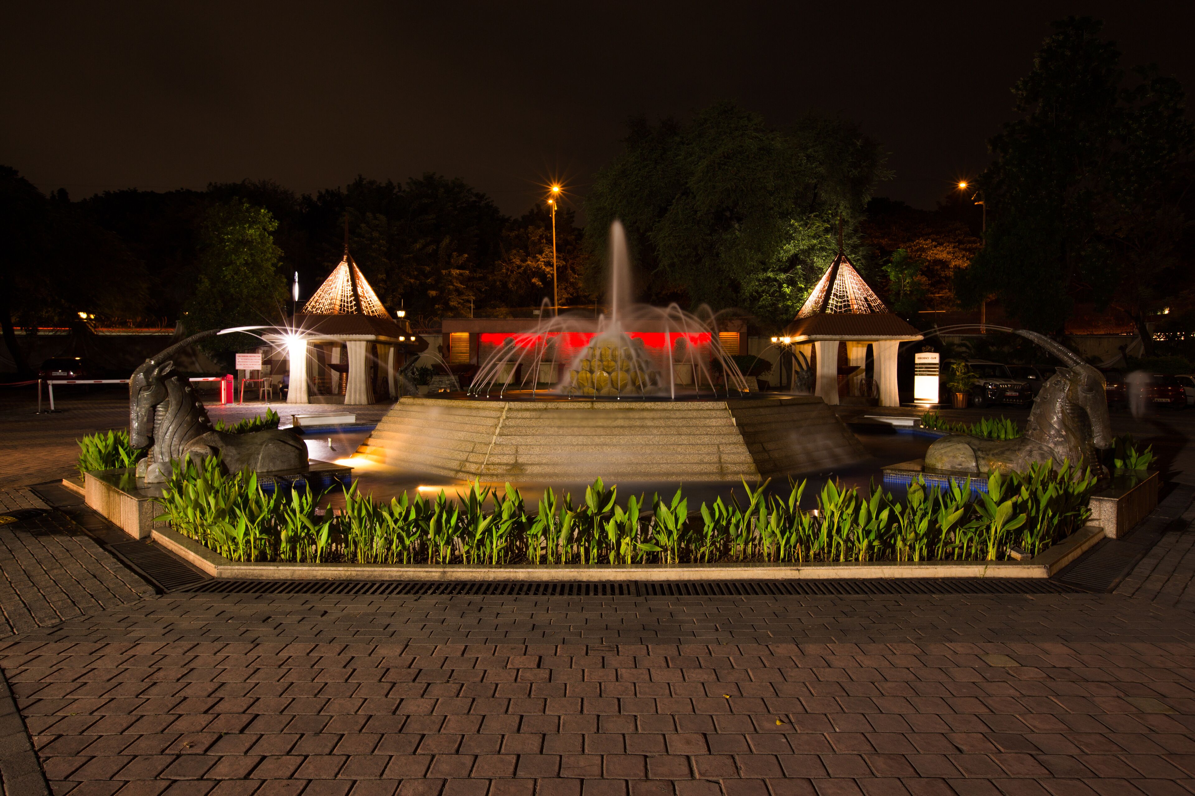 Une vue nocturne d’une fontaine bien éclairée, flanquée de deux petits pavillons illuminés de chaque côté, le tout entouré d’un chemin en briques et de verdure.