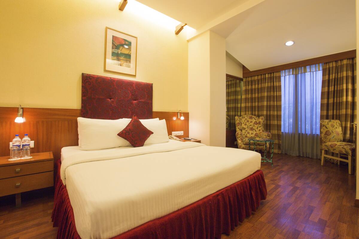 A hotel room with a large bed, wooden headboard, two bedside tables with lamps, a small table with chairs, and a window with curtains.