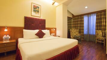 Habitación de hotel con una cama grande, cabecera de madera, dos mesitas de noche con lámparas, una pequeña mesa con sillas y una ventana con cortinas.