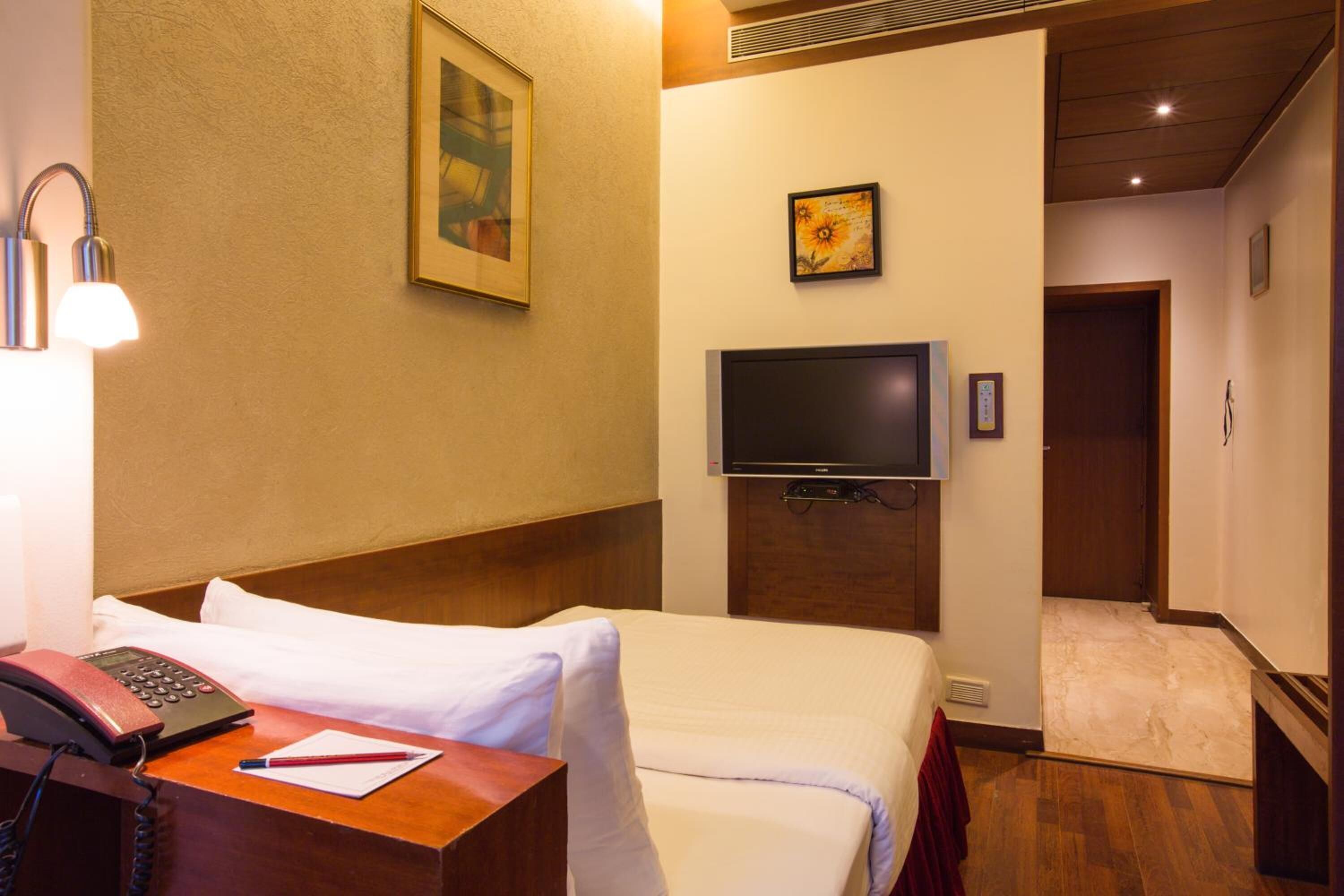 A hotel room with a bed, a desk with a telephone and a pen, a television mounted on the wall, and a door leading to another room.
