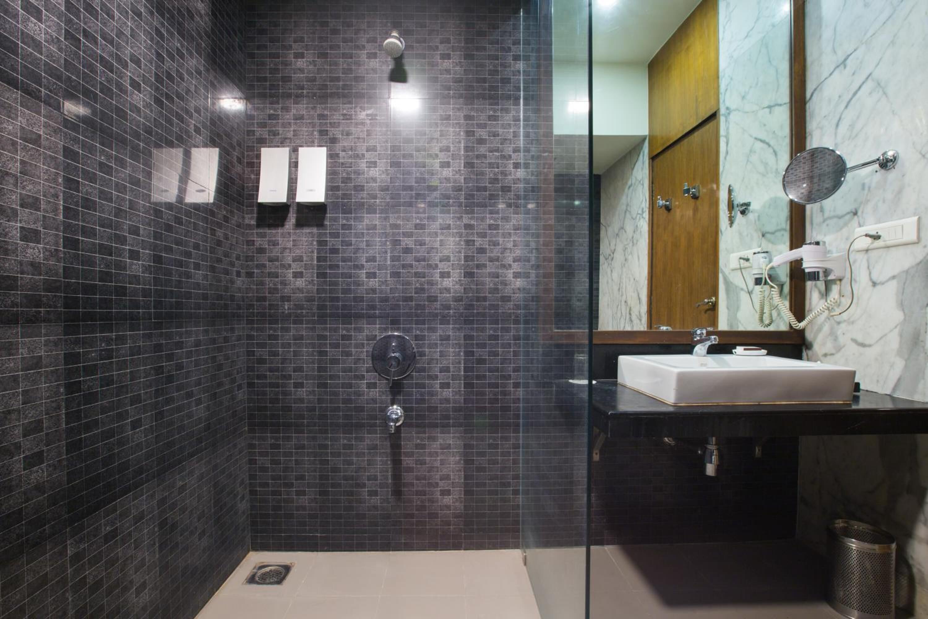 Un baño moderno con paredes de azulejos oscuros, cabina de ducha de vidrio, lavamanos blanco y tocador con encimera de mármol.