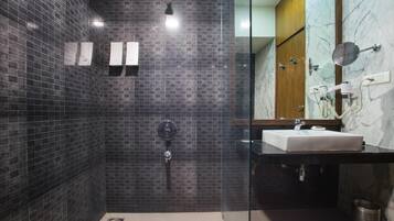 Un baño moderno con paredes de azulejos oscuros, cabina de ducha de vidrio, lavamanos blanco y tocador con encimera de mármol.