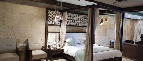 A modern hotel room with a four-poster bed, a wooden nightstand, a seating area with two chairs, and a decorative wall panel.